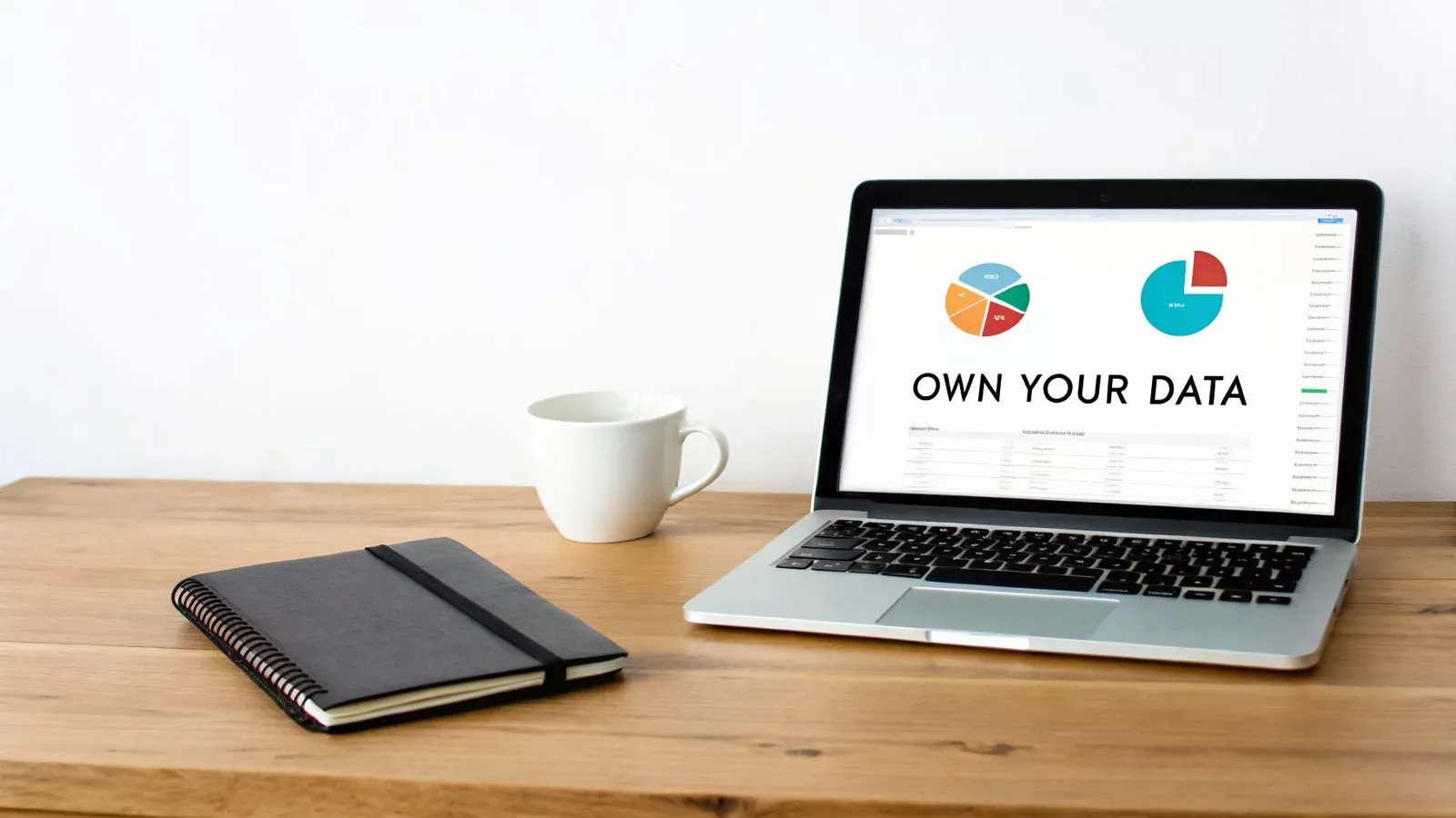 A laptop displaying 'OWN YOUR DATA' with pie charts, a notebook, and a coffee mug on a wooden desk.